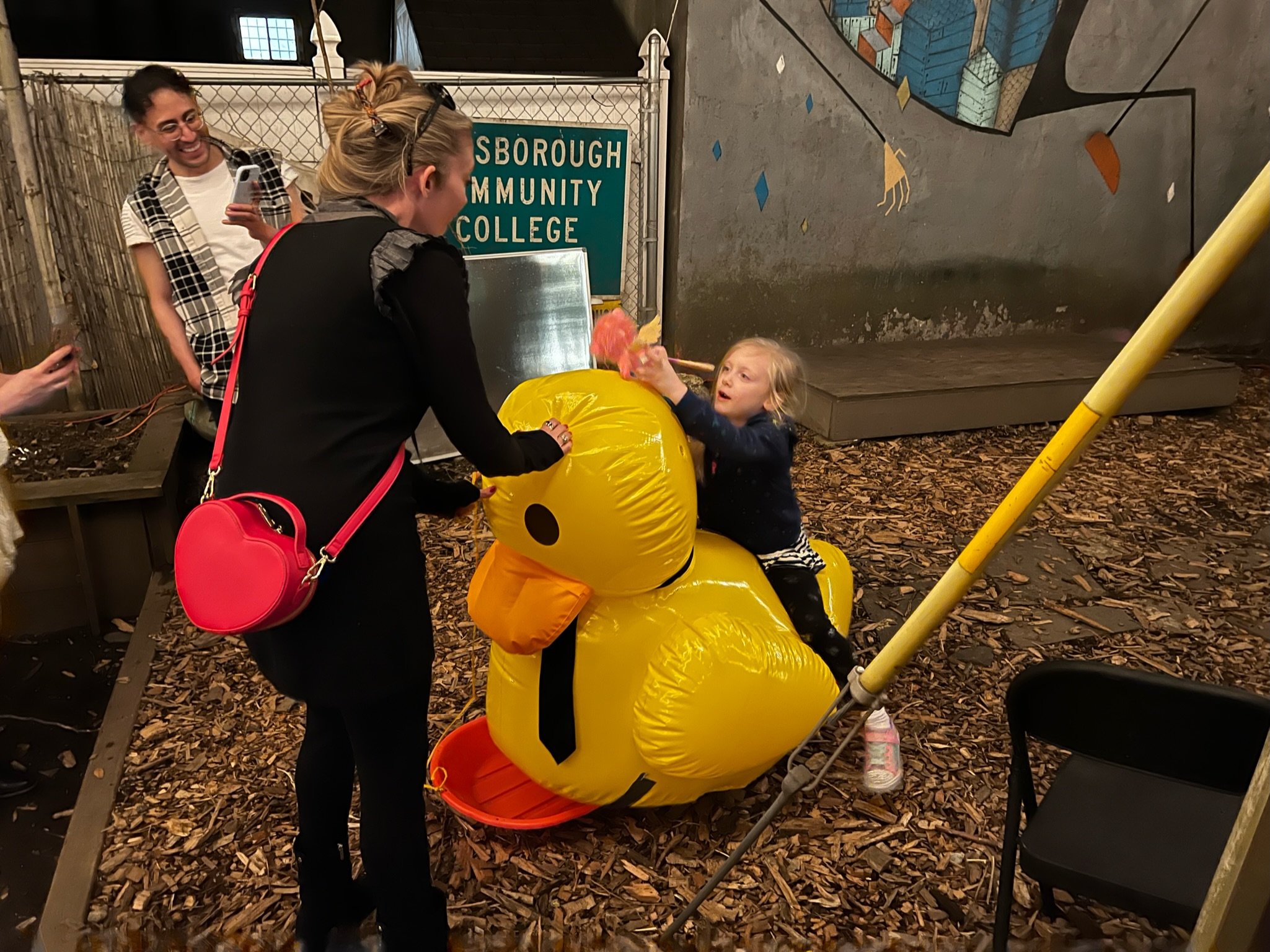 a little girl sits on a large inflatable duck, stabilized by his kind caretaker